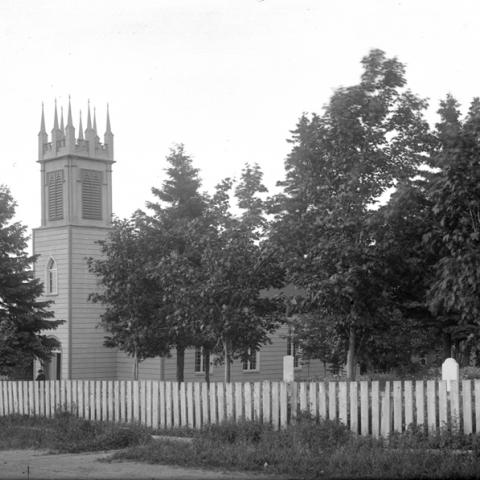 Vue sur une petite église au clocher de style néo-gothique, avec le cimetière attenant. Le terrain est fermé par une clôture.