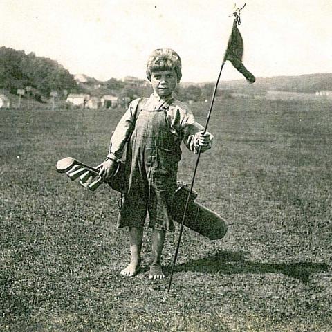 Un enfant caddie, sac de golf à l’épaule, tient un drapeau. Il est pieds nus.